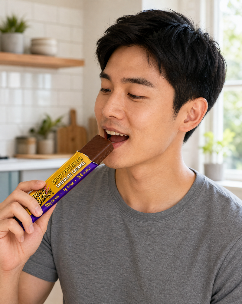 A man in a kitchen biting into a chocolate caramel bar with the wrapper peeled down and still held in the same hand