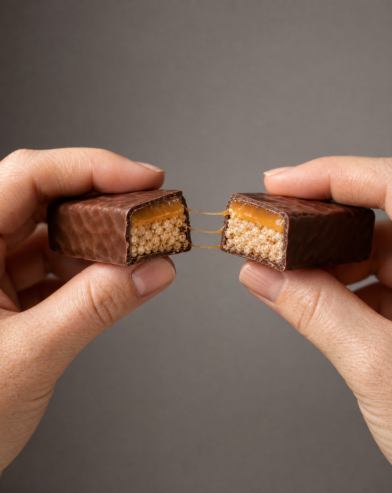 Two hands pulling a chocolate caramel bar apart, caramel stretching into short strings between the halves with the crisp layer visible in both cross-sections