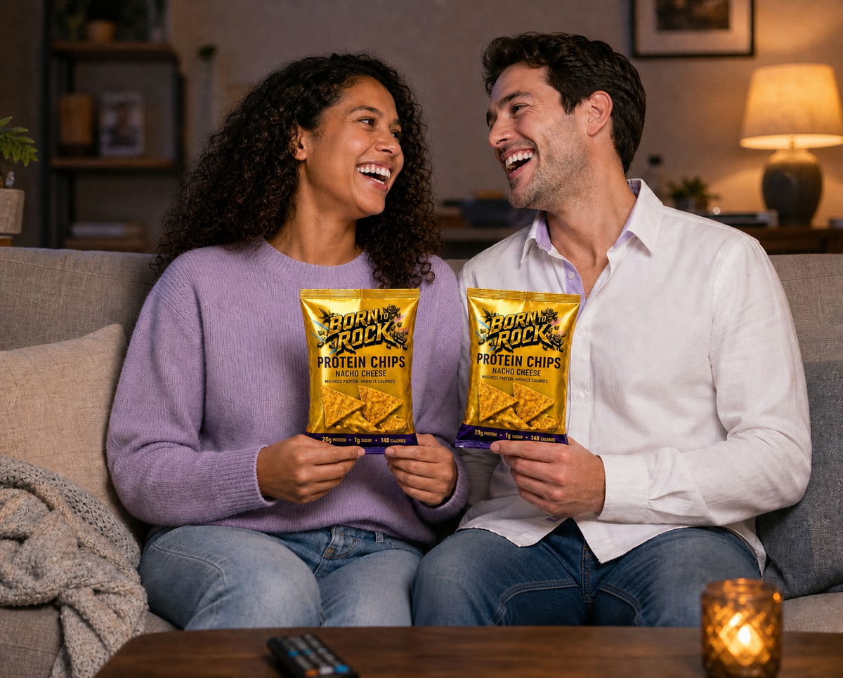 Two people laughing on a sofa in the evening, each holding a bag of nacho protein chips