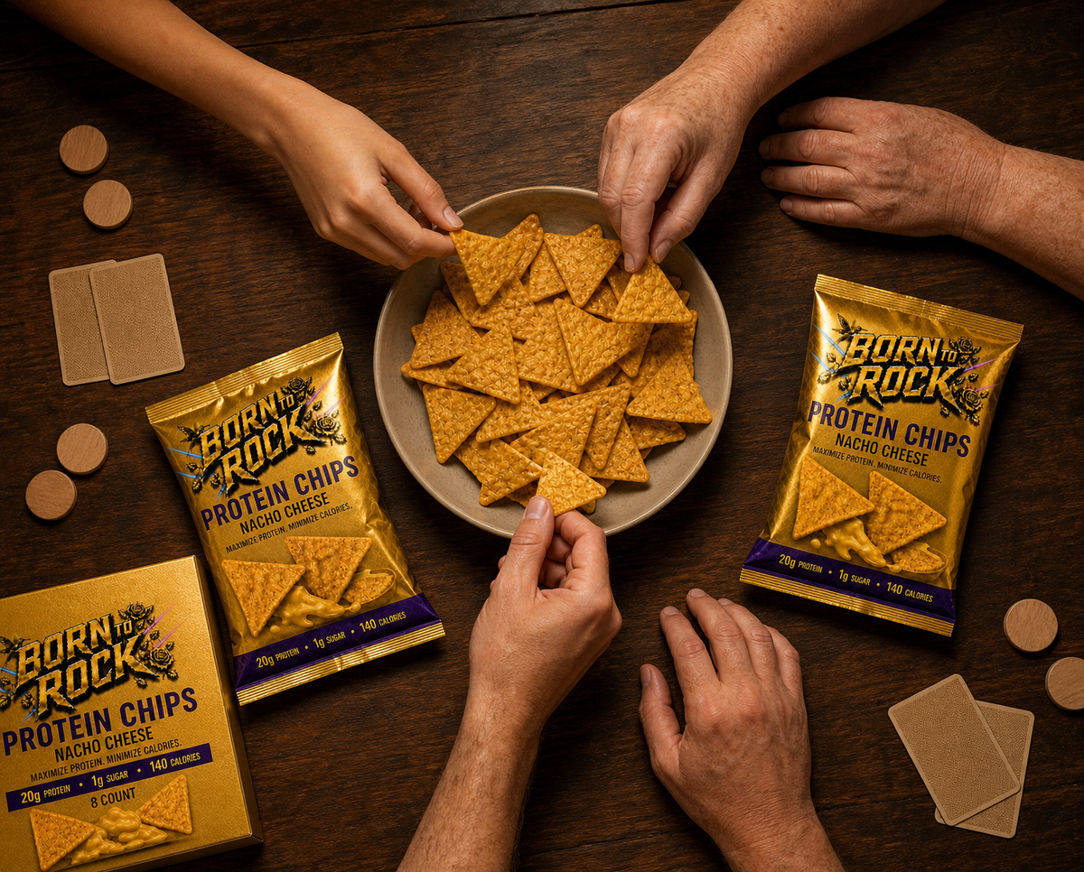 Overhead view of a game night table, three pairs of hands reaching into a bowl of nacho protein chips beside two open bags and the display box